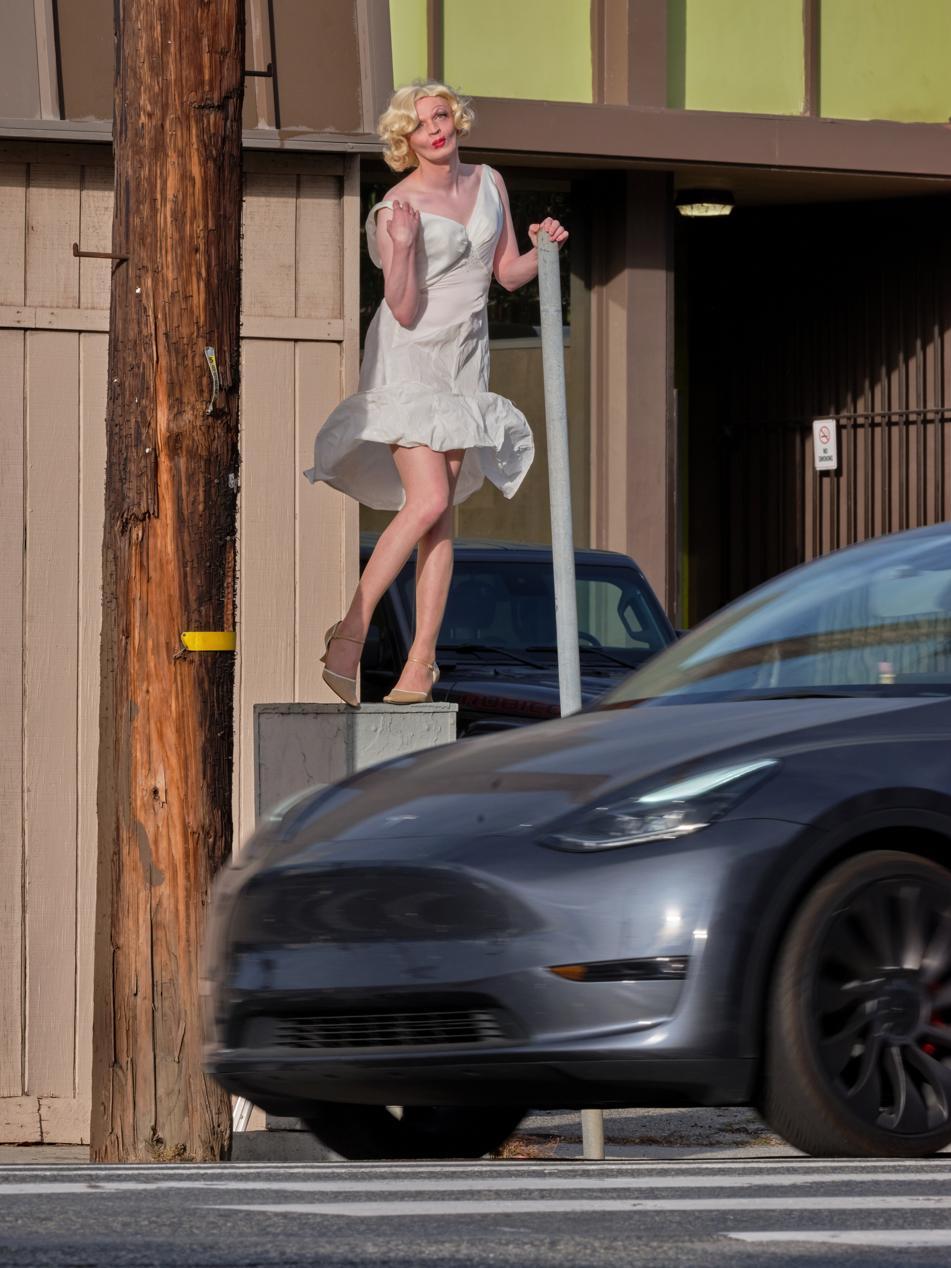 Brooks Ginnan dressed as marilyn monore with her dress flying up in the wind standing on an electrical box coyly while traffic passes by
